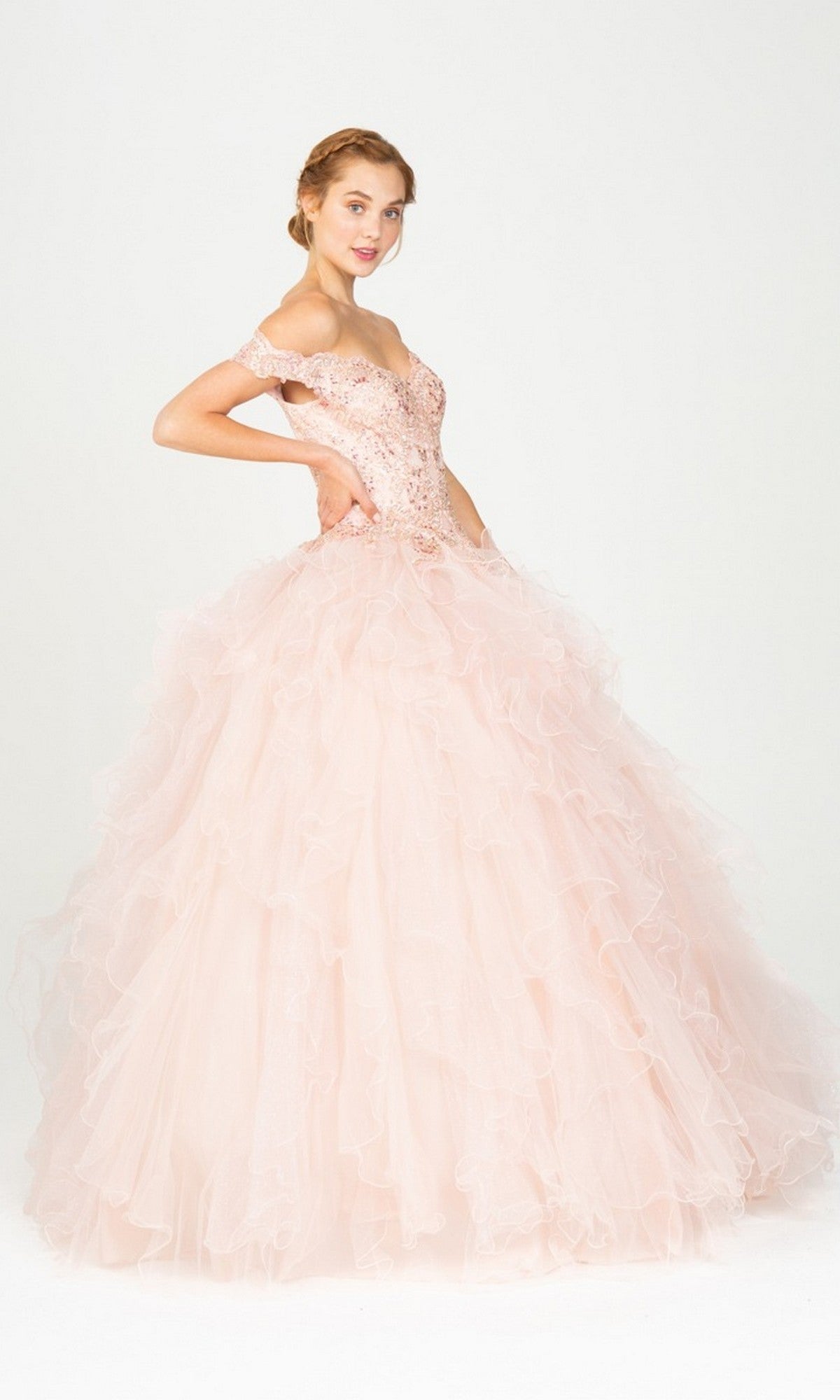 A woman poses in the Quinceanera Dress 9313 by Eureka, showcasing its blush pink hue, beaded off-the-shoulder bodice, and full ruffled tulle skirt for a stunning ball gown look against a plain white background.